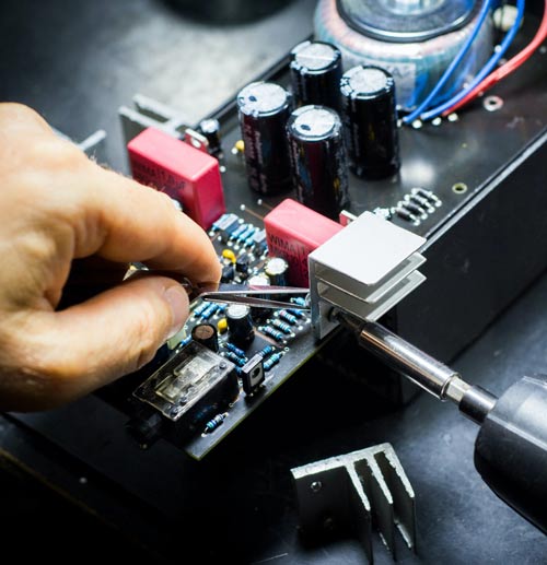 A hand using tweezers to work on a circuit board with various components, including capacitors and heat sinks, in a home electronics repair setting.