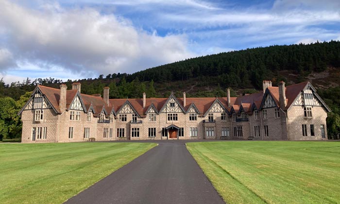 a large, historic stone mansion with a symmetrical facade, set against a green hill and blue sky, featuring a long driveway and manicured lawns