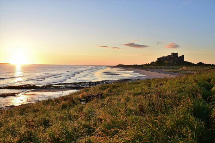 A scenic view of a sandy beach at sunset, with gentle waves lapping at the shore and lush green grass in the foreground, featuring a historic castle on a distant hill.