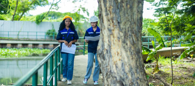 Two women walking beside a tree on a pathway near water, one wearing a hard hat and the other wearing a helmet, both holding documents in a green and lush outdoor environment.