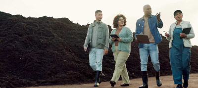 A group of four people, dressed in work attire and boots, walking together and discussing near a large pile of compost or soil.