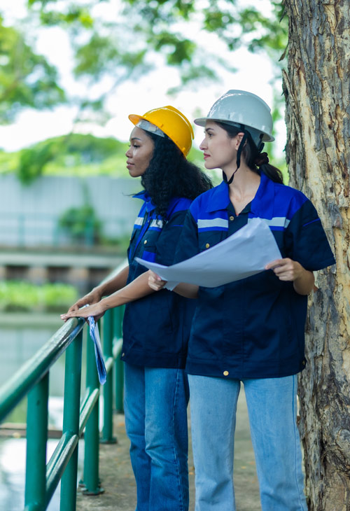 Two women in safety helmets, one orange and one white, are standing by a railing near a tree, examining blueprints, showcasing teamwork in a construction or engineering setting.