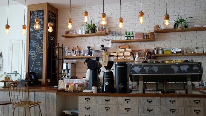 Cozy coffee shop interior with a wooden counter, espresso machine, and pendant lighting, featuring shelves stocked with products and decor.