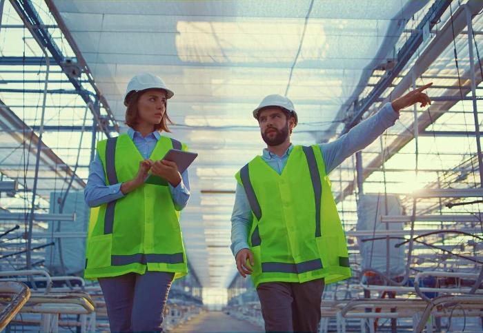 Two construction workers wearing safety helmets and bright green vests are walking through a large industrial space, discussing plans as one points forward, presumably reviewing the engineering or architecture of the site.